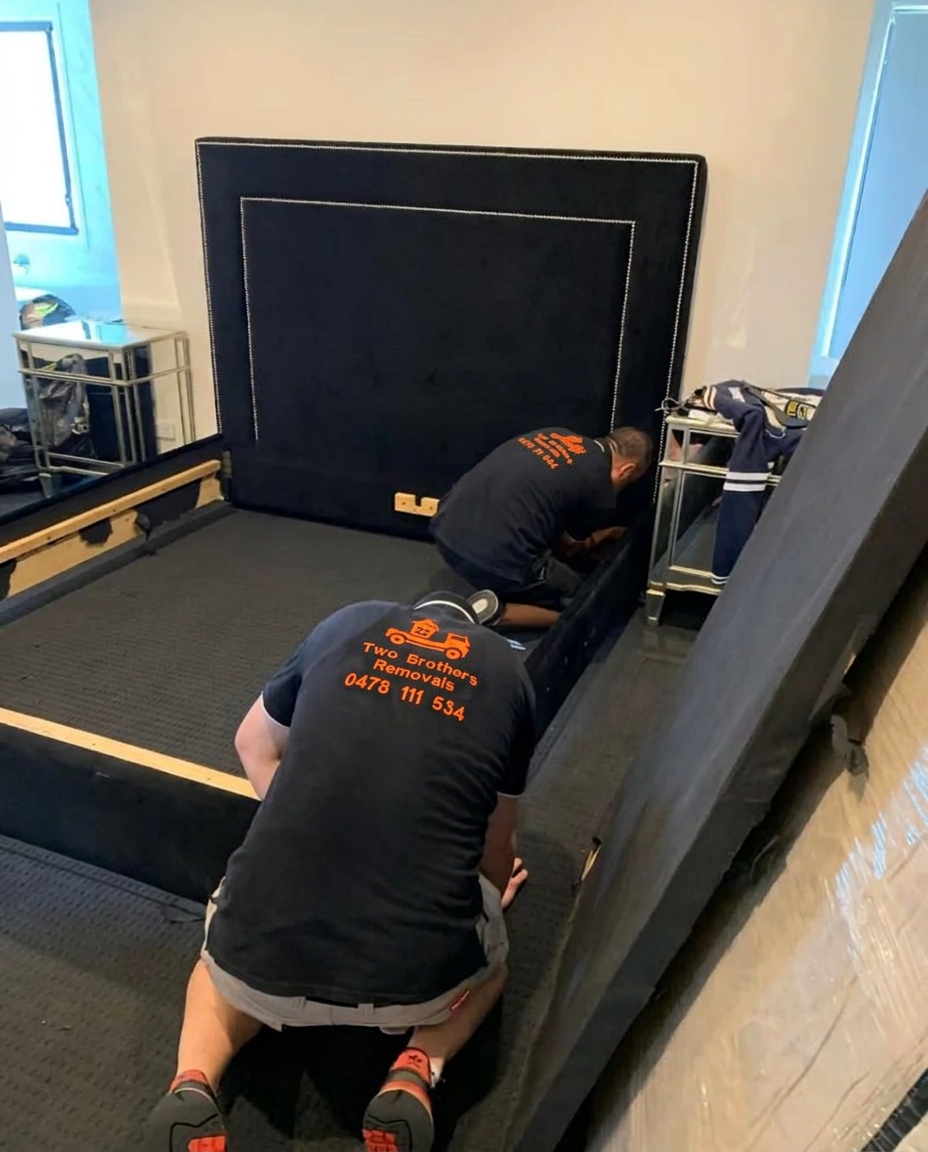 Two removalists carefully dismantling an upholstered bed frame and headboard