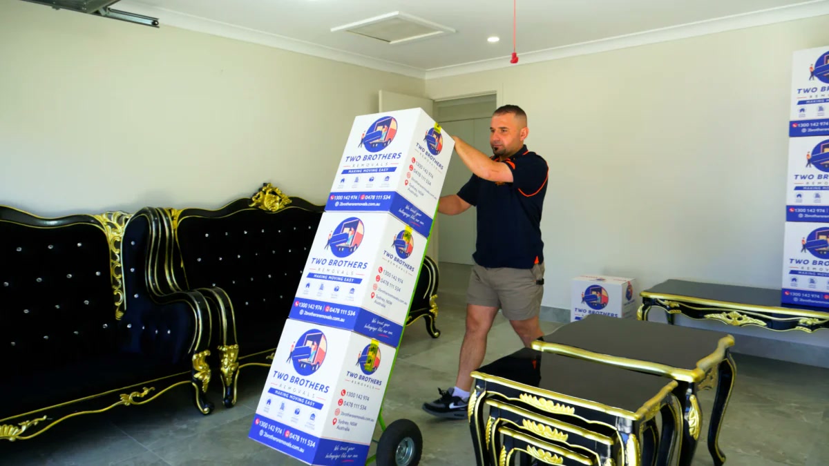 Removalist wheeling stacked Two Brothers boxes on a hand trolley through the living room